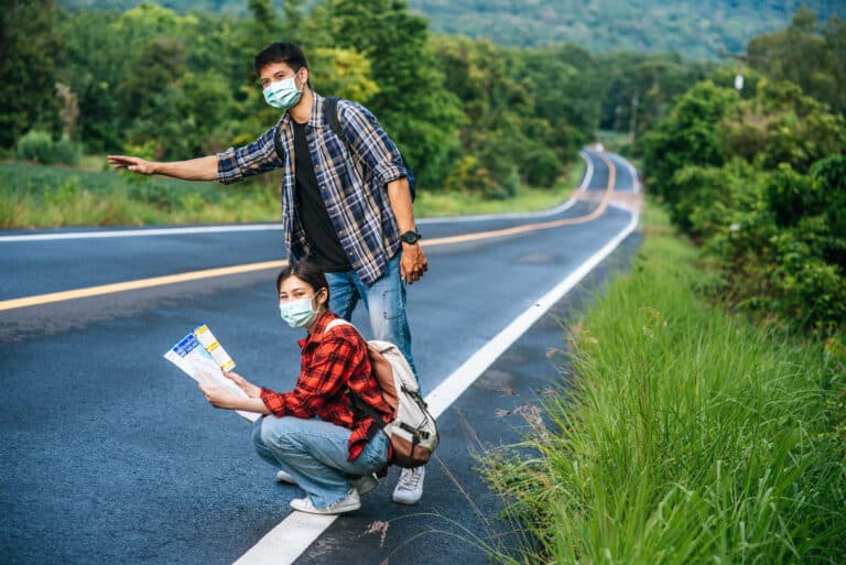 Notre Blog 6 Female tourists sit and look at the map, male tourists pretending to hitchhike. Both wear masks and are on the side of the road.